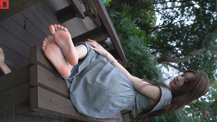 A pure and beautiful Chinese girl named Yang Yang, wearing a blue long dress, playfully showcases her fair and tender feet on a wooden table in the park