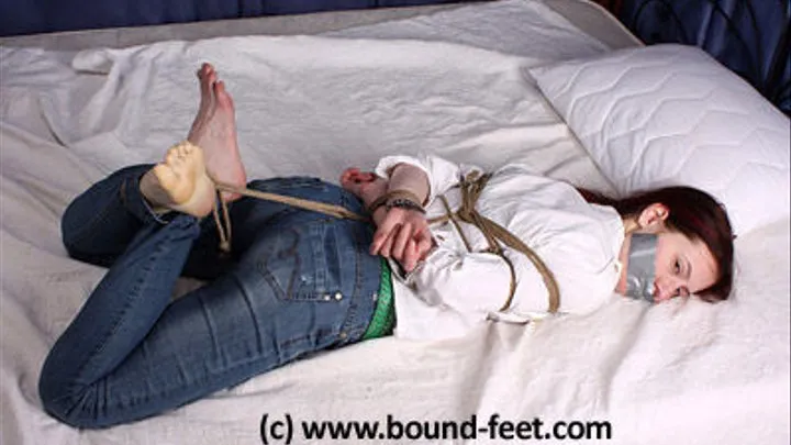 Patricia hogtied on the bed wearing jeans and shirt, barefoot, tape-gagged