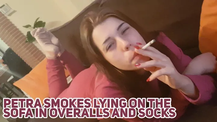 Petra smokes lying on the sofa in overalls and socks